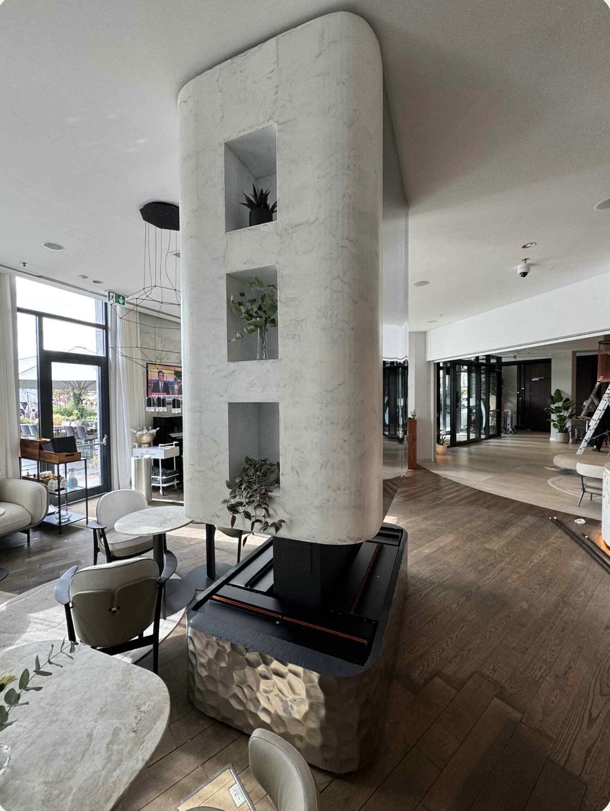 Marmurowa kolumna z wnękami na rośliny w nowoczesnym wnętrzu biurowym w Toruniu. Widoczne stoliki i krzesła w strefie relaksu, drewniana podłoga i duże okna.
