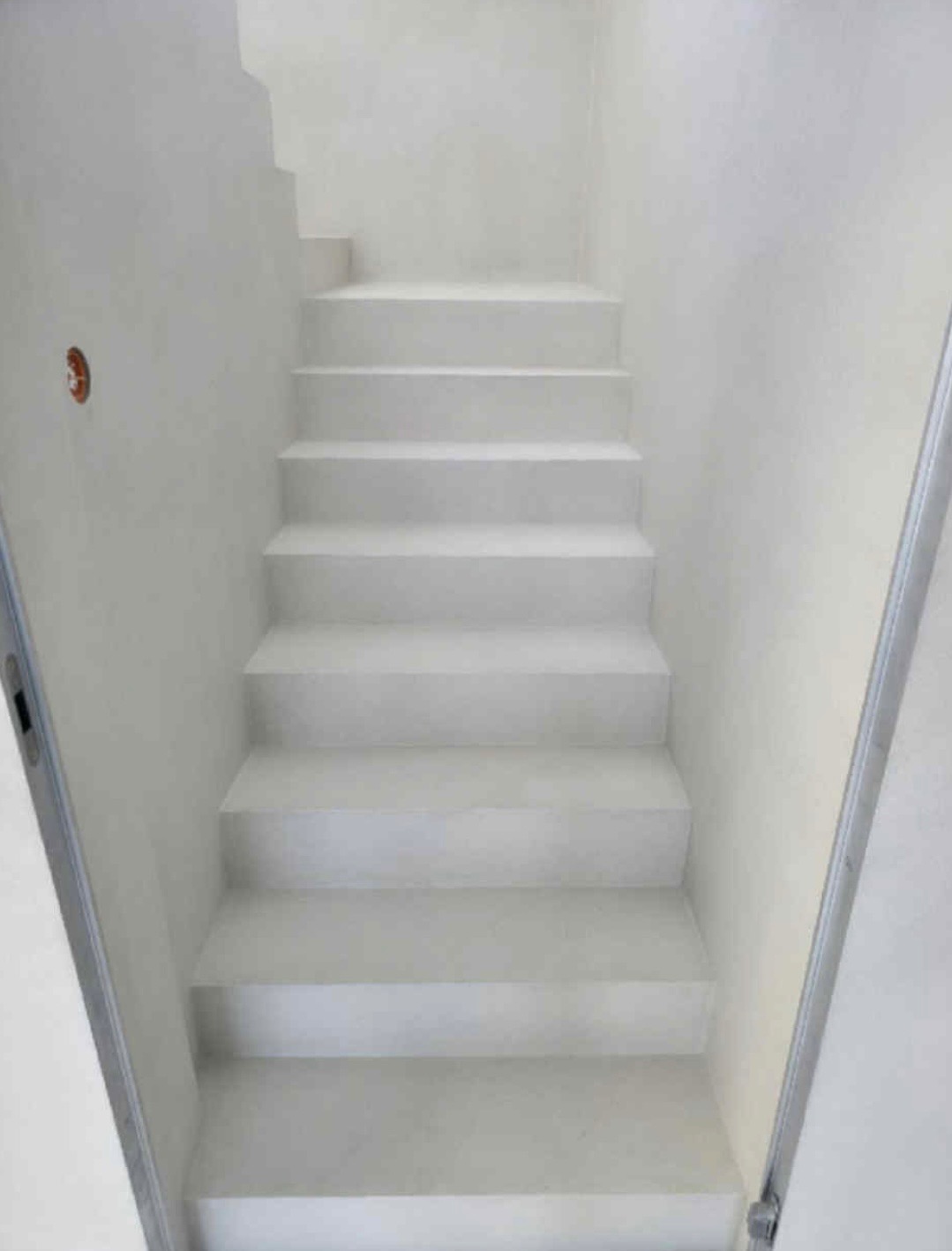 Białe schody w minimalistycznym wnętrzu, ściany i stopnie w jednolitej tonacji, widoczny fragment instalacji elektrycznej i metalowa krawędź drzwi.