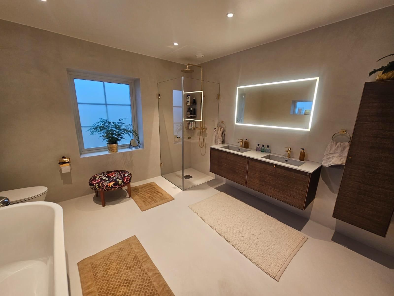 Nowoczesna łazienka z kabiną prysznicową, podwójną umywalką z szafką z ciemnego drewna, podświetlanym lustrem i dekoracyjnym stołkiem obok okna.