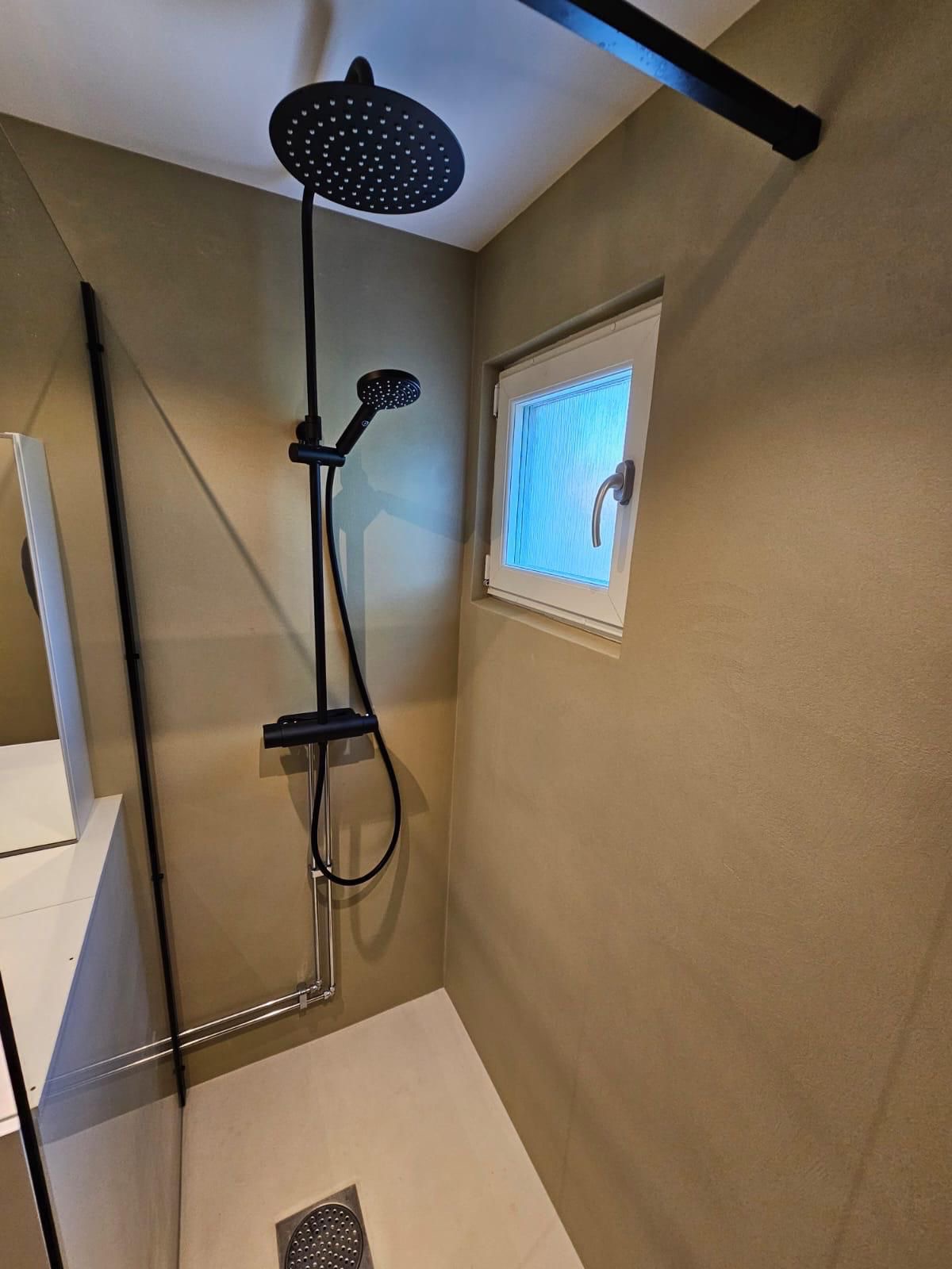 Nowoczesna kabina prysznicowa z czarną armaturą i deszczownicą w łazience o beżowych ścianach. Minimalistyczny design z oknem i odpływem liniowym.