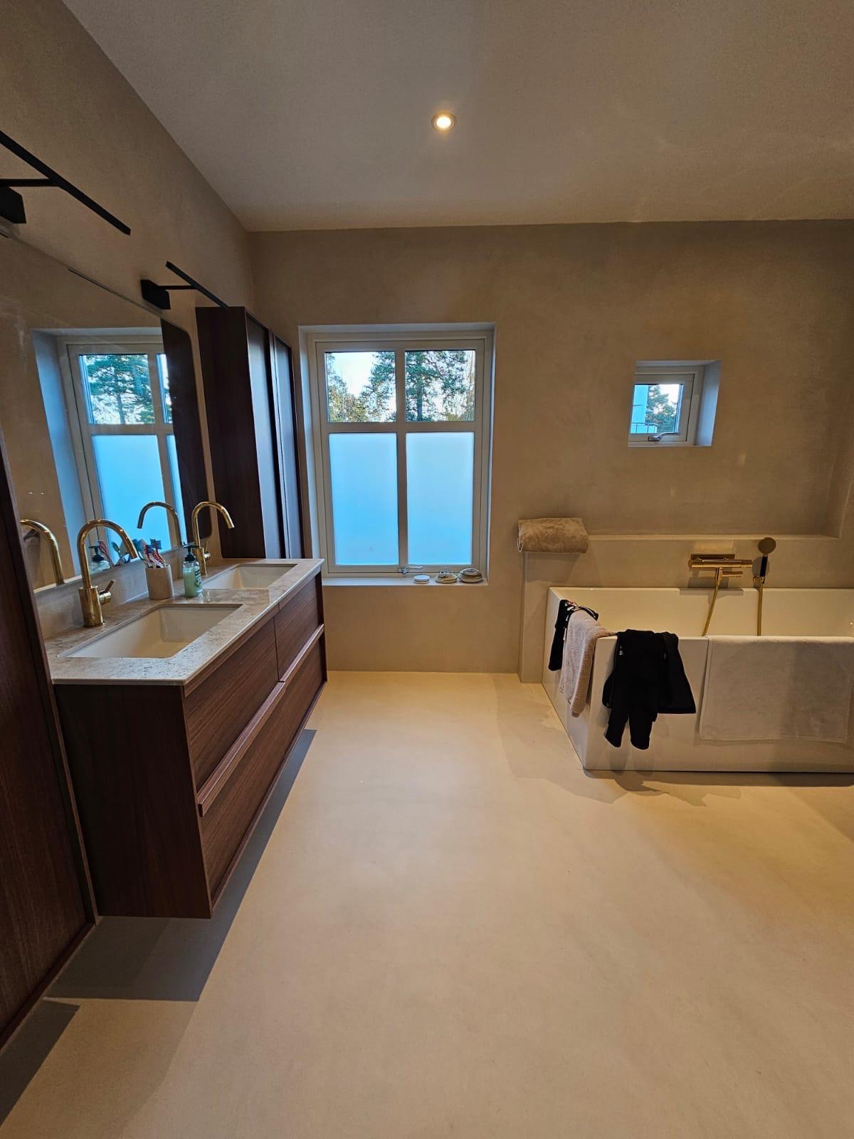 Nowoczesna łazienka z podwójną umywalką na drewnianej szafce, wolnostojącą wanną i oknami z matową folią. Minimalistyczny design, beżowe ściany i podłoga.