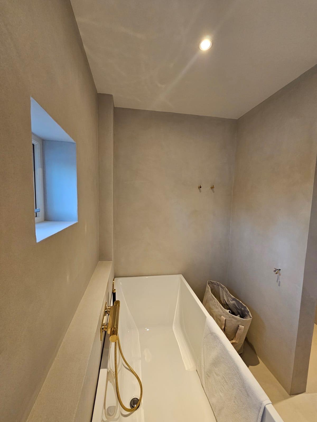 Minimalistyczna łazienka po remoncie: biała wanna z złotą armaturą, beżowe ściany z fakturą, małe okno. Nowoczesny design, stonowana kolorystyka.