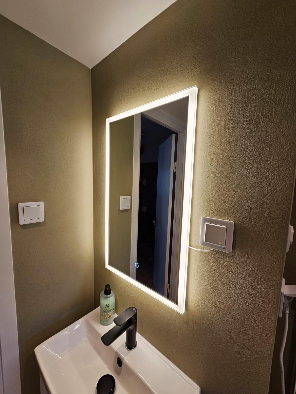 Nowoczesna łazienka z podświetlanym lustrem, umywalką i czarną baterią. Ściany w odcieniach beżu z widoczną fakturą. Minimalistyczny design i ciepłe oświetlenie.