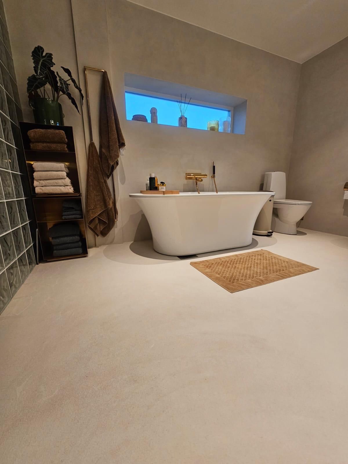 Nowoczesna łazienka w beżowych tonacjach z wolnostojącą wanną, złotą armaturą i minimalistycznym oknem. Na podłodze beżowy dywanik, a obok wanny toaleta.