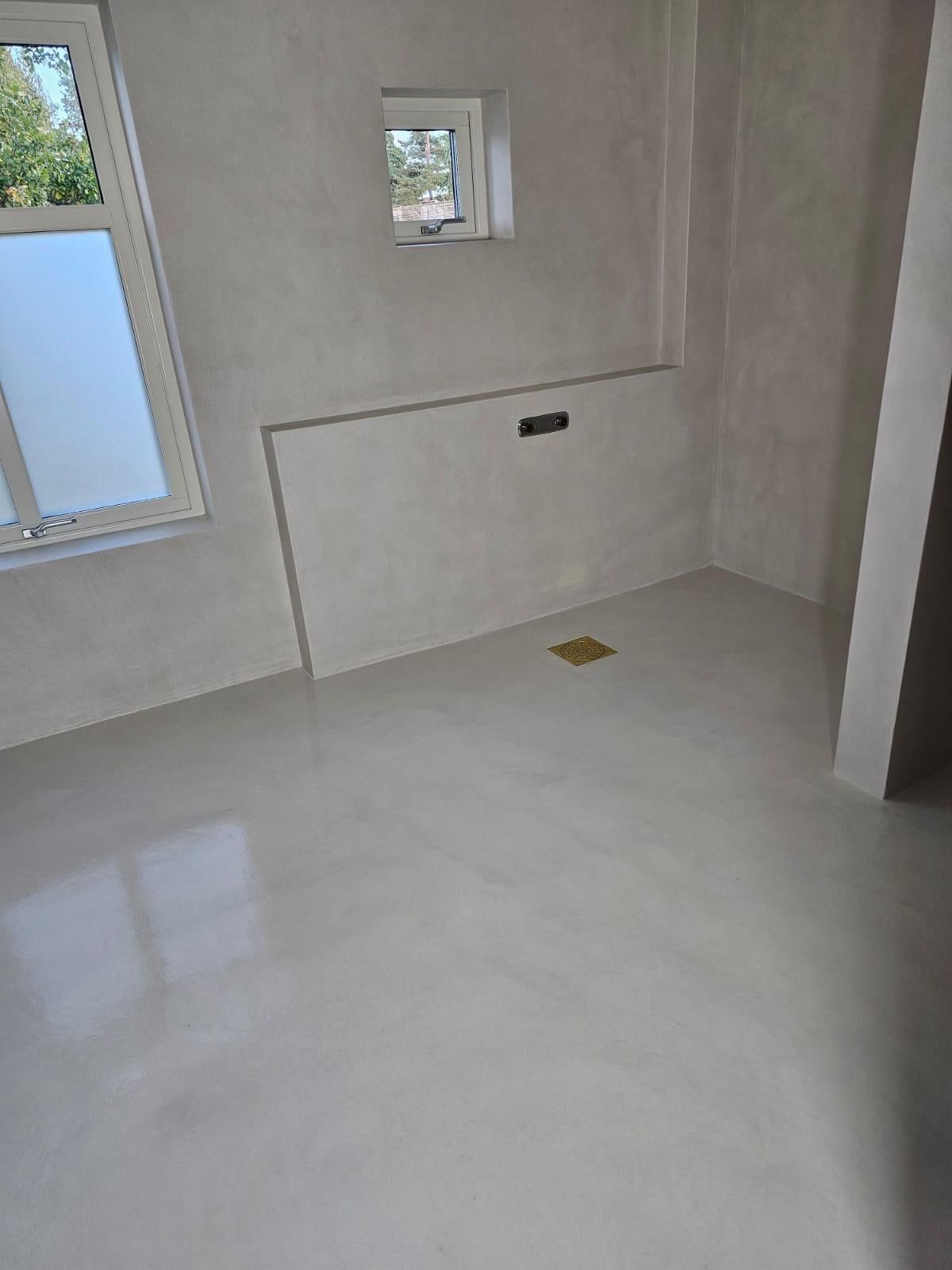 Minimalistyczna łazienka po remoncie: gładkie, szare ściany i podłoga z odpływem liniowym. Widoczne okno z mleczną szybą i małe okienko wentylacyjne.