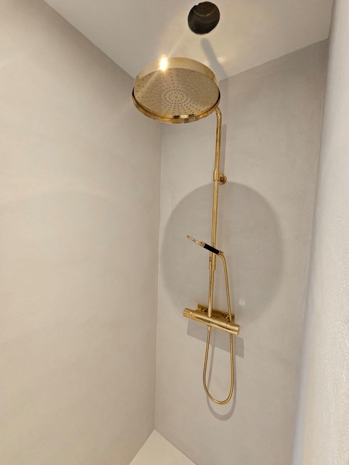 Złota deszczownica z termostatem na tle jasnych ścian w wykończonej łazience. Widoczny otwór w suficie. Minimalistyczny design.