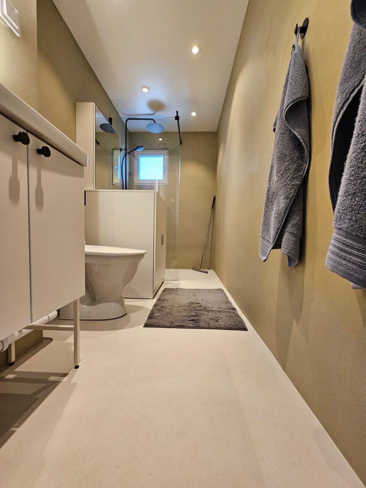 Nowoczesna łazienka z kabiną prysznicową typu walk-in, umywalką z szafką, toaletą i szarym dywanikiem. Na ścianie wiszą dwa szare ręczniki.