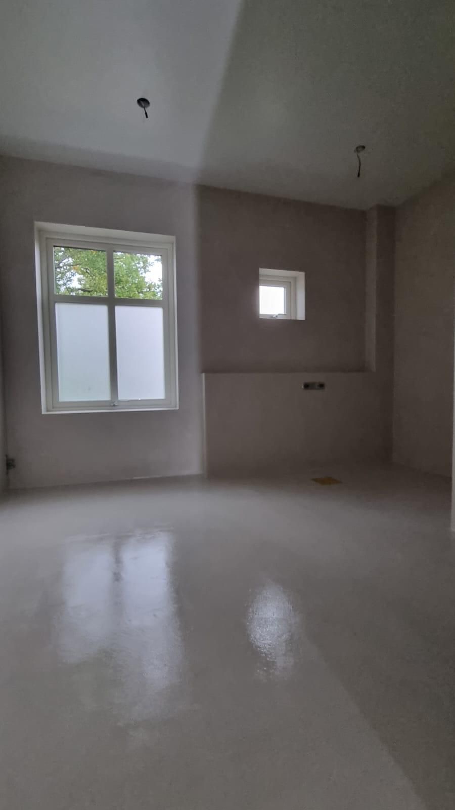 Wnętrze po szpachlowaniu: gładkie, jasne ściany, okna z widokiem na zieleń. Przygotowane do dalszych prac wykończeniowych, widoczne punkty oświetleniowe na suficie.