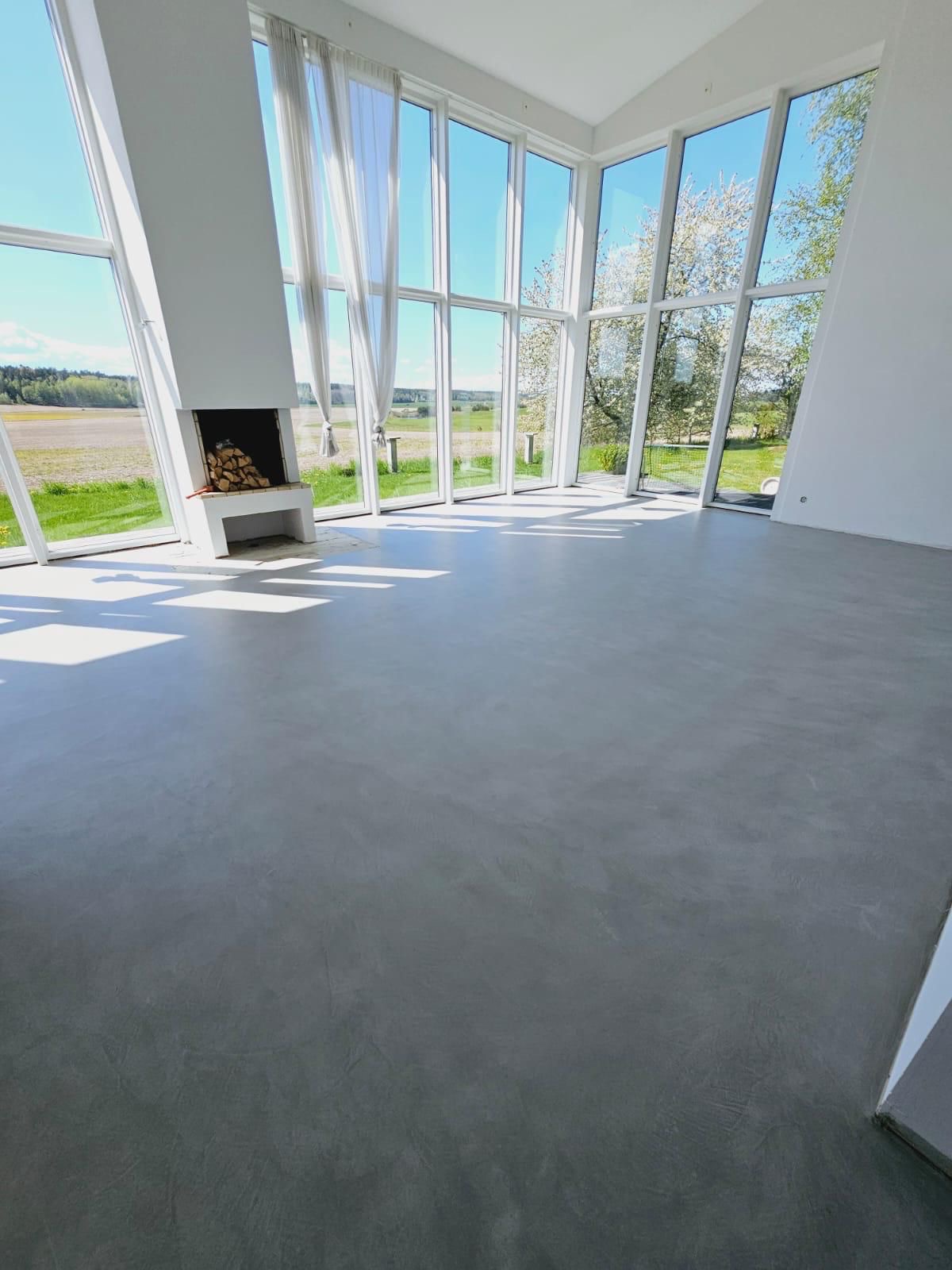 Jasne wnętrze z betonową posadzką, dużymi oknami z widokiem na zieleń i kominkiem z drewnem. Minimalistyczny design, nowoczesne wykończenie, dużo naturalnego światła.