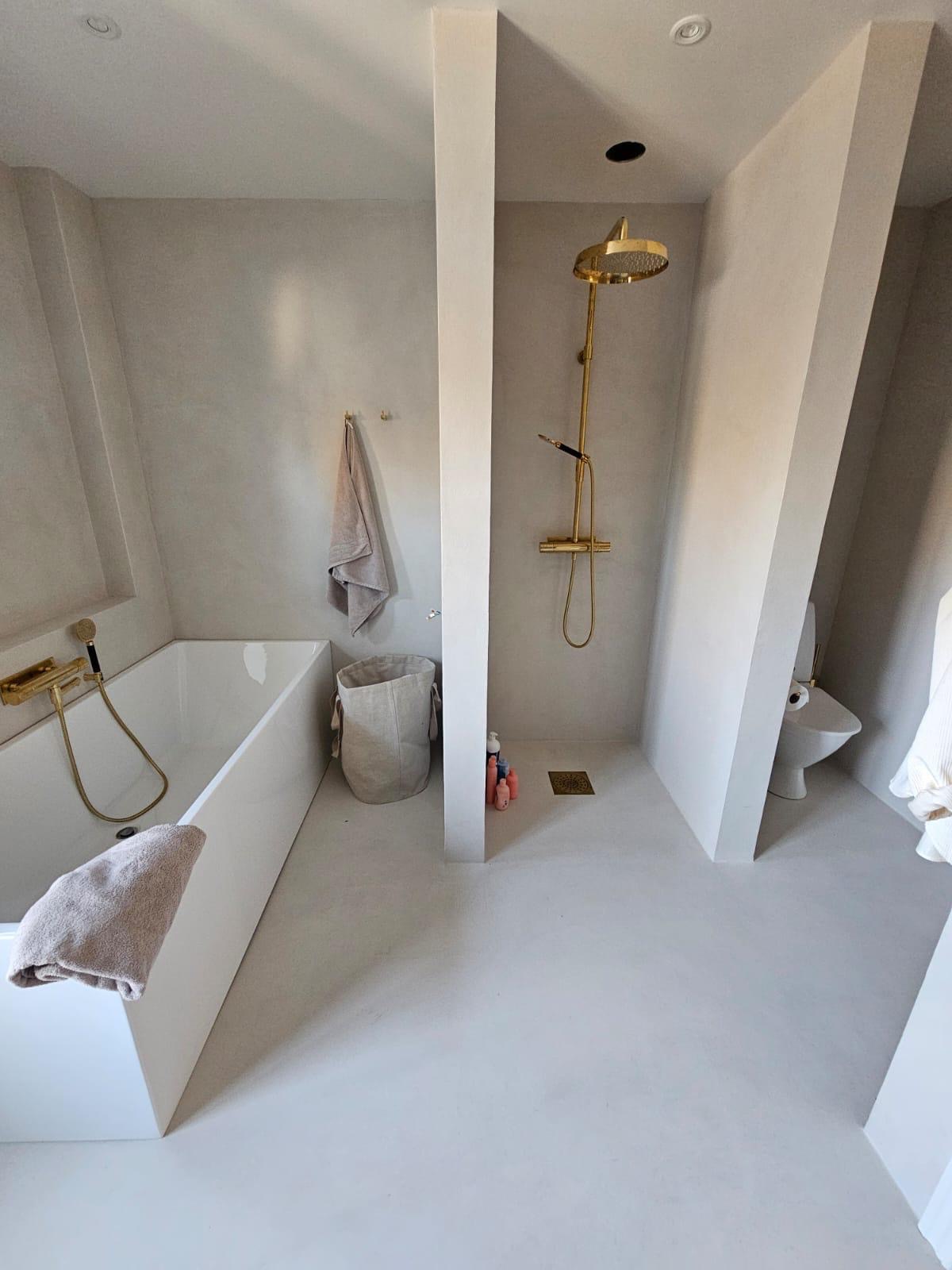 Minimalistyczna łazienka z beżowymi ścianami, złotą armaturą i kabiną prysznicową oddzieloną ścianką. Widoczna wanna, toaleta i kosz na pranie.