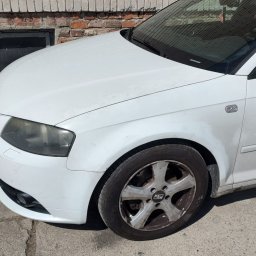 FENIX CENTRUM - Biały samochód Audi A3 z brudną felgą stoi na tle ceglanej ściany. Widoczny fragment przedniego błotnika, reflektora i koła. Auto zaparkowane na betonowym podłożu.