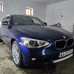 FENIX CENTRUM - Błyszczący, granatowy BMW serii 1 po detailingu, widoczne odbicia świateł na lakierze, wnętrze warsztatu z oknami w tle, tablica rejestracyjna WA 0559K.