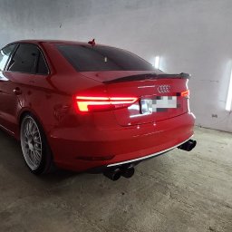 FENIX CENTRUM - Czerwone Audi A3 z tuningowanym spojlerem i czterema końcówkami wydechu, sfotografowane w warsztacie pod kątem, z włączonymi tylnymi światłami.