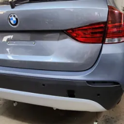 Szary samochód BMW w warsztacie, widok z tyłu, z widocznym spawarką i butlą z gazem w tle. Detale karoserii i wyposażenie warsztatu.