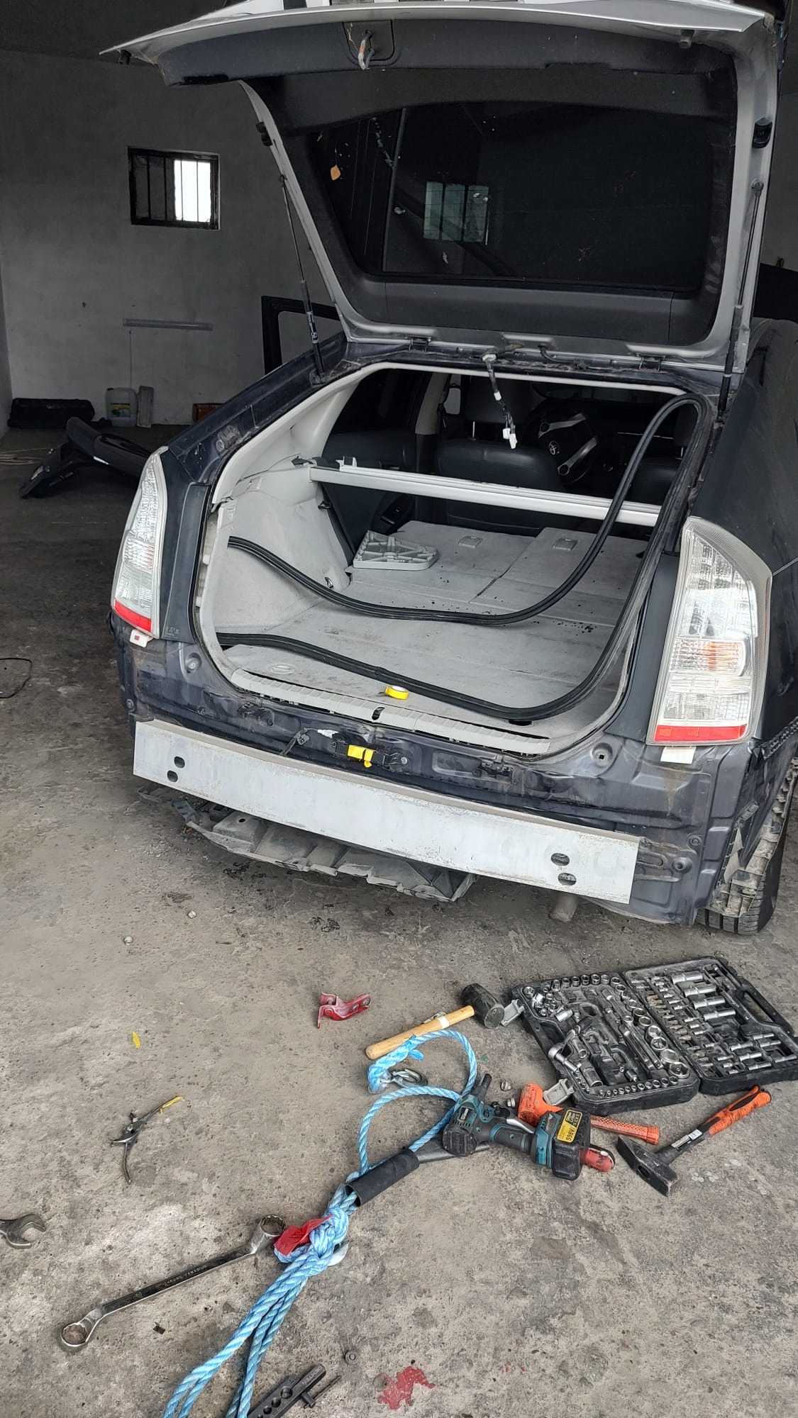 Uszkodzony tył auta (Prius) w warsztacie, z otwartą klapą bagażnika, narzędzia na podłodze. Widoczny nowy, surowy element karoserii przed montażem.
