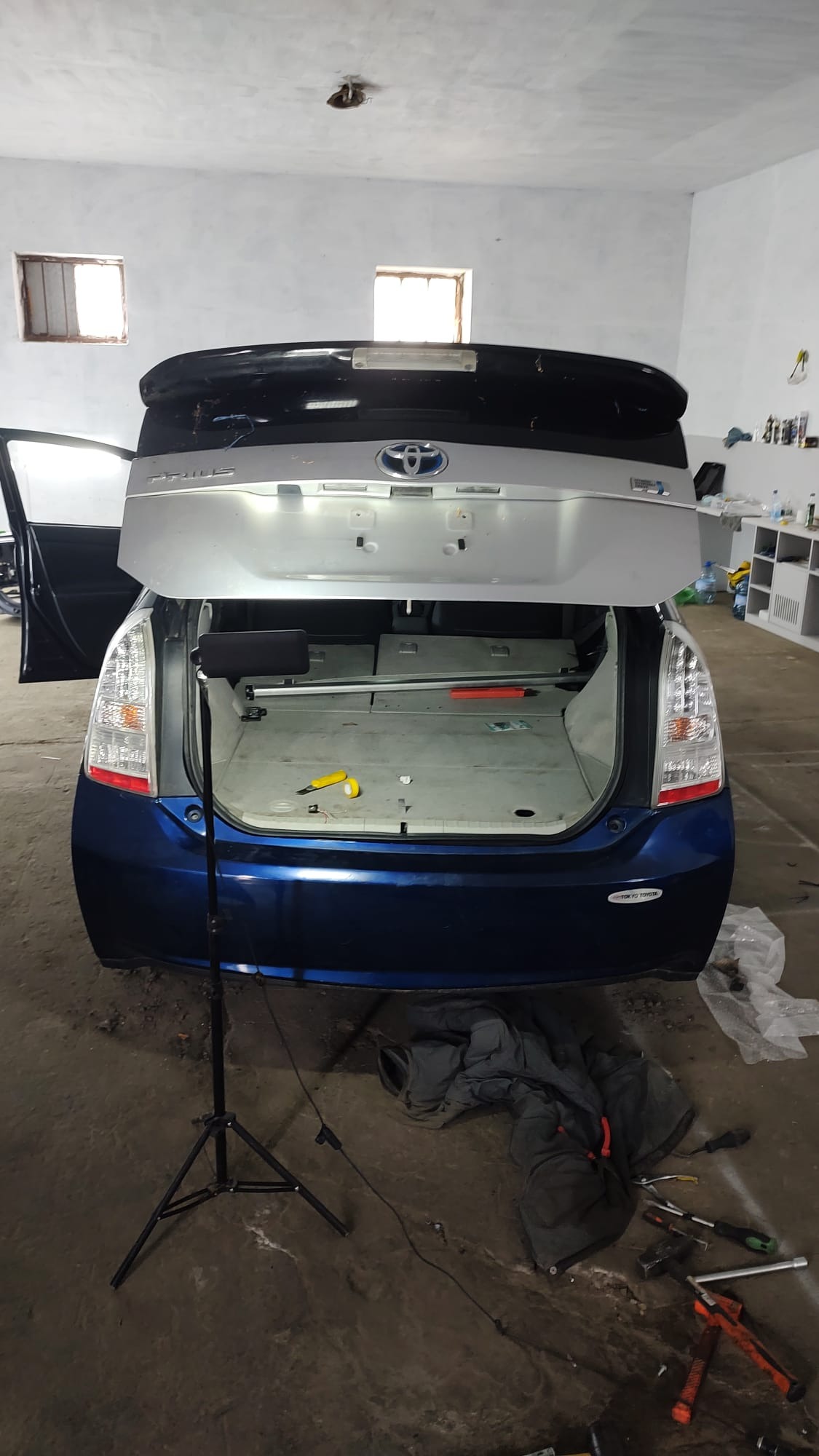 Błękitny Toyota Prius z otwartym bagażnikiem w warsztacie. Widoczne narzędzia, lampa oświetleniowa i elementy wnętrza bagażnika w trakcie demontażu.