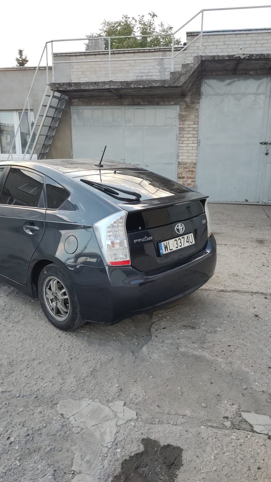 Czarny samochód Toyota Prius, widok z tyłu, zaparkowany przed garażem z szarymi drzwiami i metalowymi schodami na zewnątrz. Tablica rejestracyjna WL 3374U.