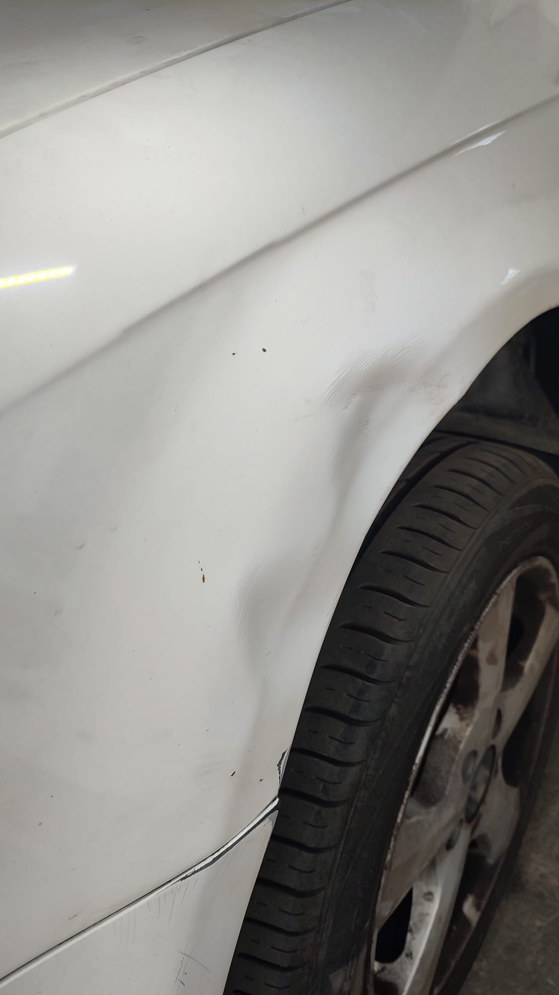 Biały samochód z wgnieceniem na błotniku przy kole. Widoczne zarysowania i zabrudzenia. Uszkodzenie karoserii.