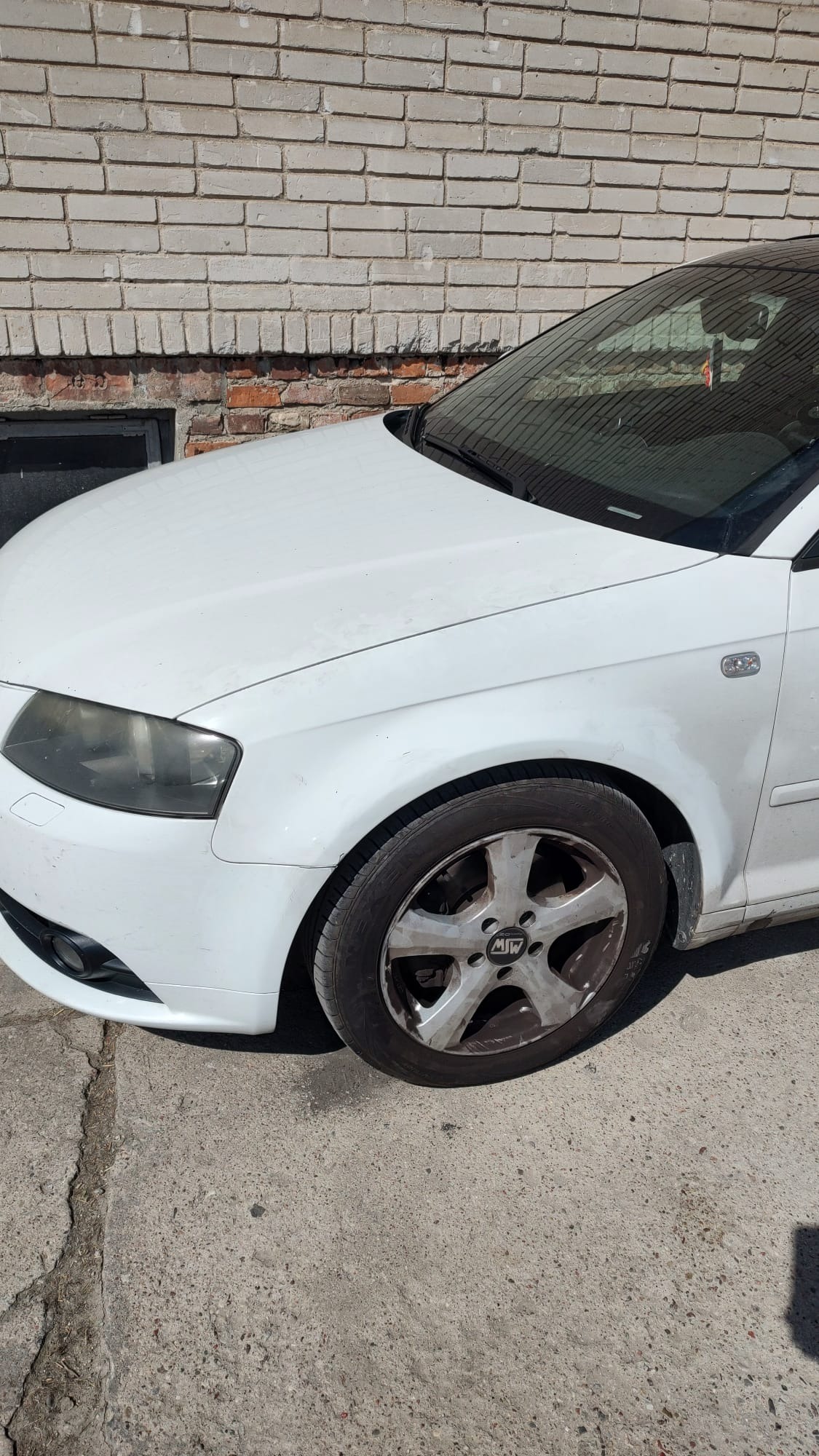 Biały samochód Audi A3 z brudną felgą stoi na tle ceglanej ściany. Widoczny fragment przedniego błotnika, reflektora i koła. Auto zaparkowane na betonowym podłożu.