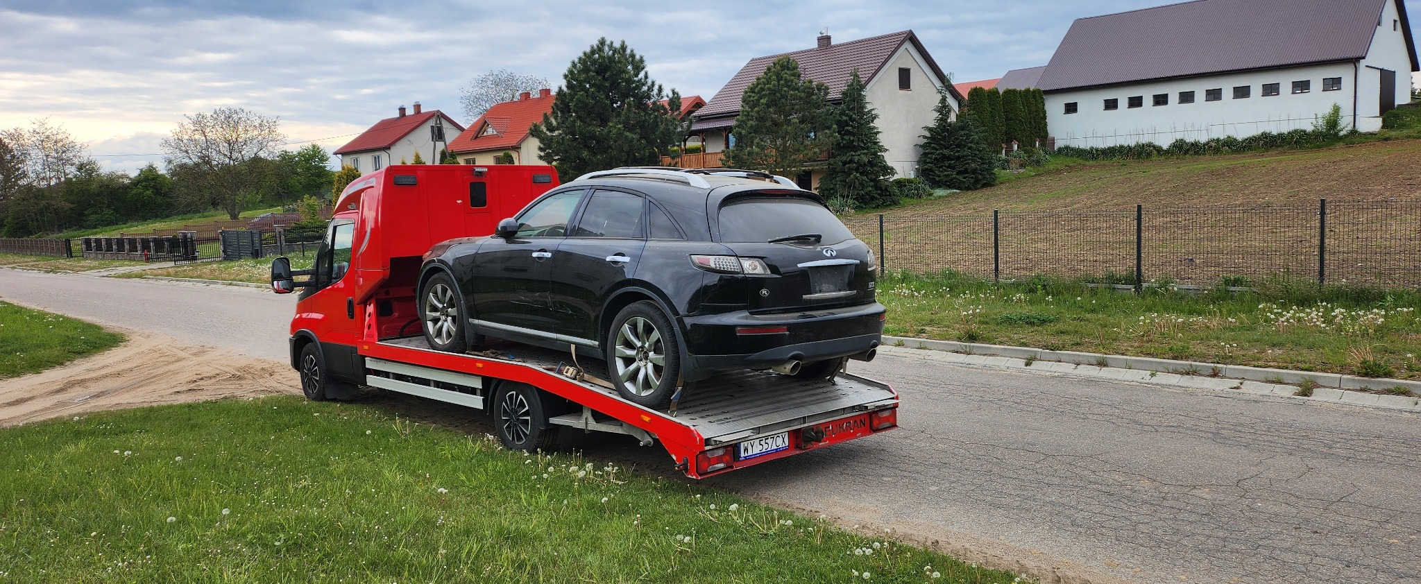 Czarny SUV Infiniti na czerwonej lawecie pomocy drogowej na tle wiejskich domów i zieleni. Widok z tyłu, tablica rejestracyjna widoczna.