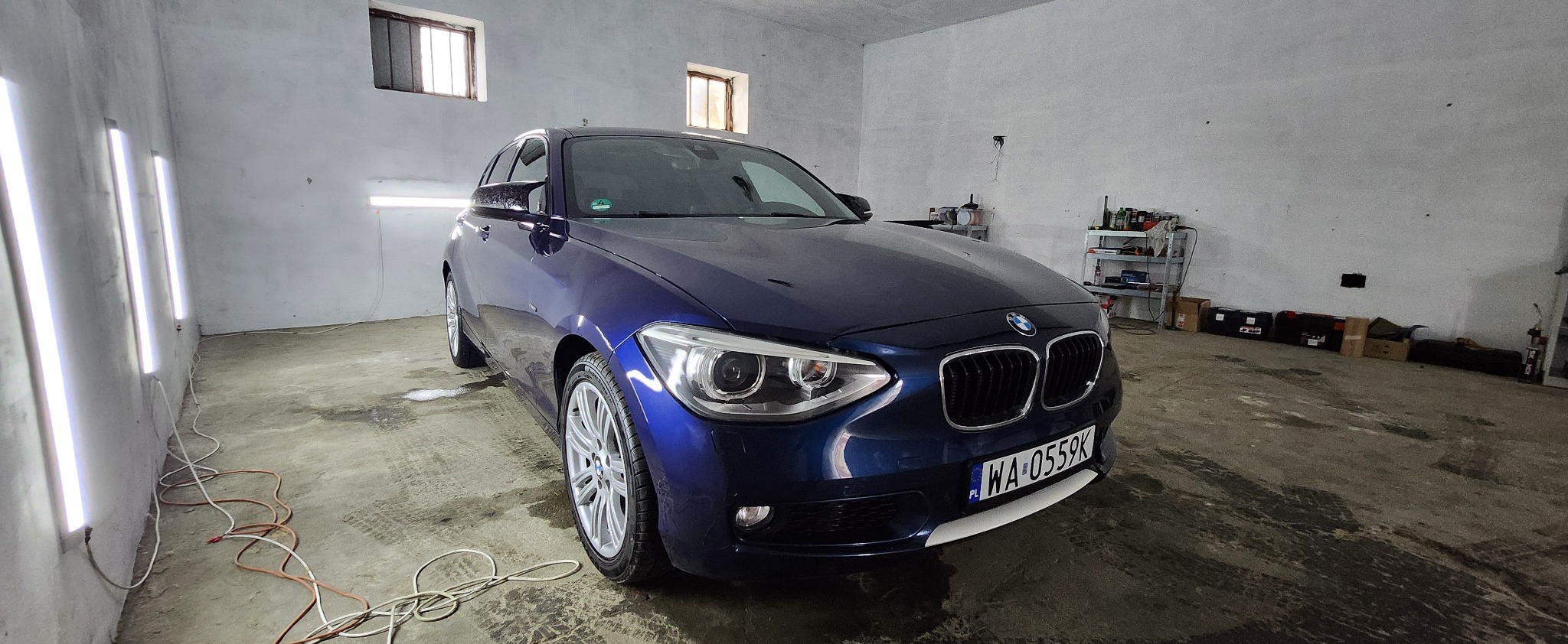 Błyszczący, granatowy BMW serii 1 po detailingu, widoczne odbicia świateł na lakierze, wnętrze warsztatu z oknami w tle, tablica rejestracyjna WA 0559K.