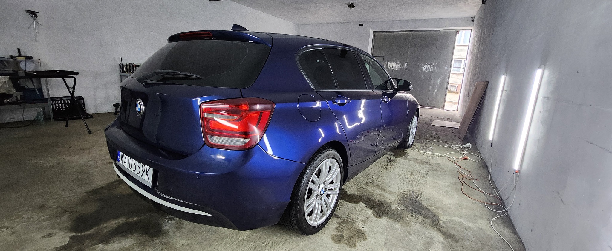 Błyszczący, granatowy BMW serii 1 po detailingu, zaparkowany w garażu z widocznymi śladami opon na posadzce. Widok z tyłu, z lekkim kątem bocznym, ukazujący odnowiony lakier.