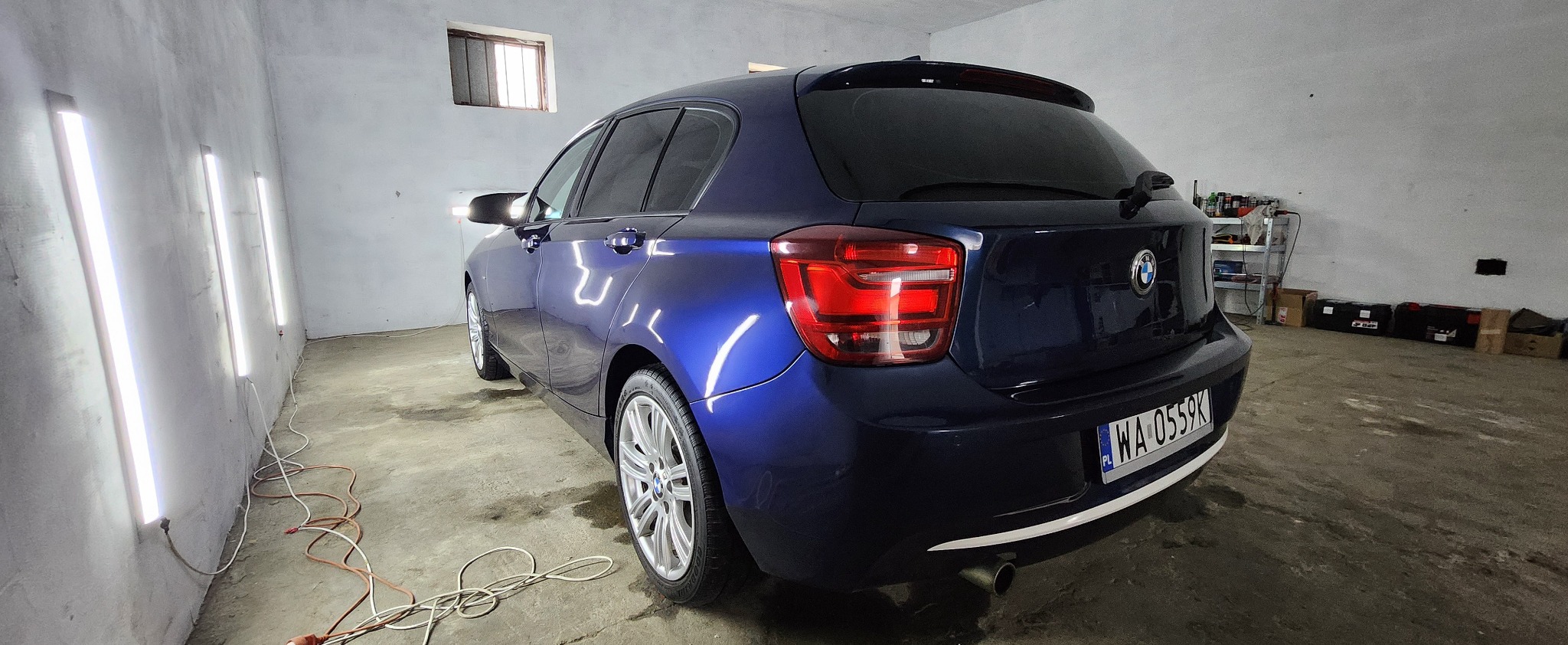 Błyszczący, granatowy BMW hatchback po detailingu w garażu z lampami inspekcyjnymi; widoczny tylny zderzak, tablica rejestracyjna i felgi aluminiowe.