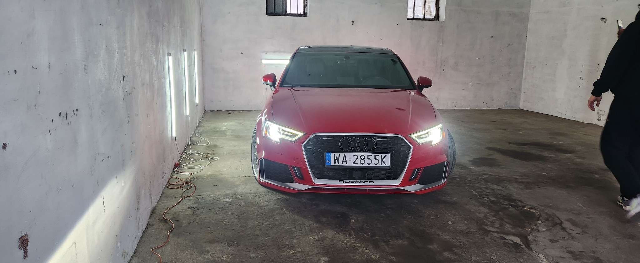 Czerwone Audi A3 Quattro z włączonymi światłami, zaparkowane w garażu z lampami inspekcyjnymi. Widoczny numer rejestracyjny WA 2855K. Garaż z surowymi ścianami.