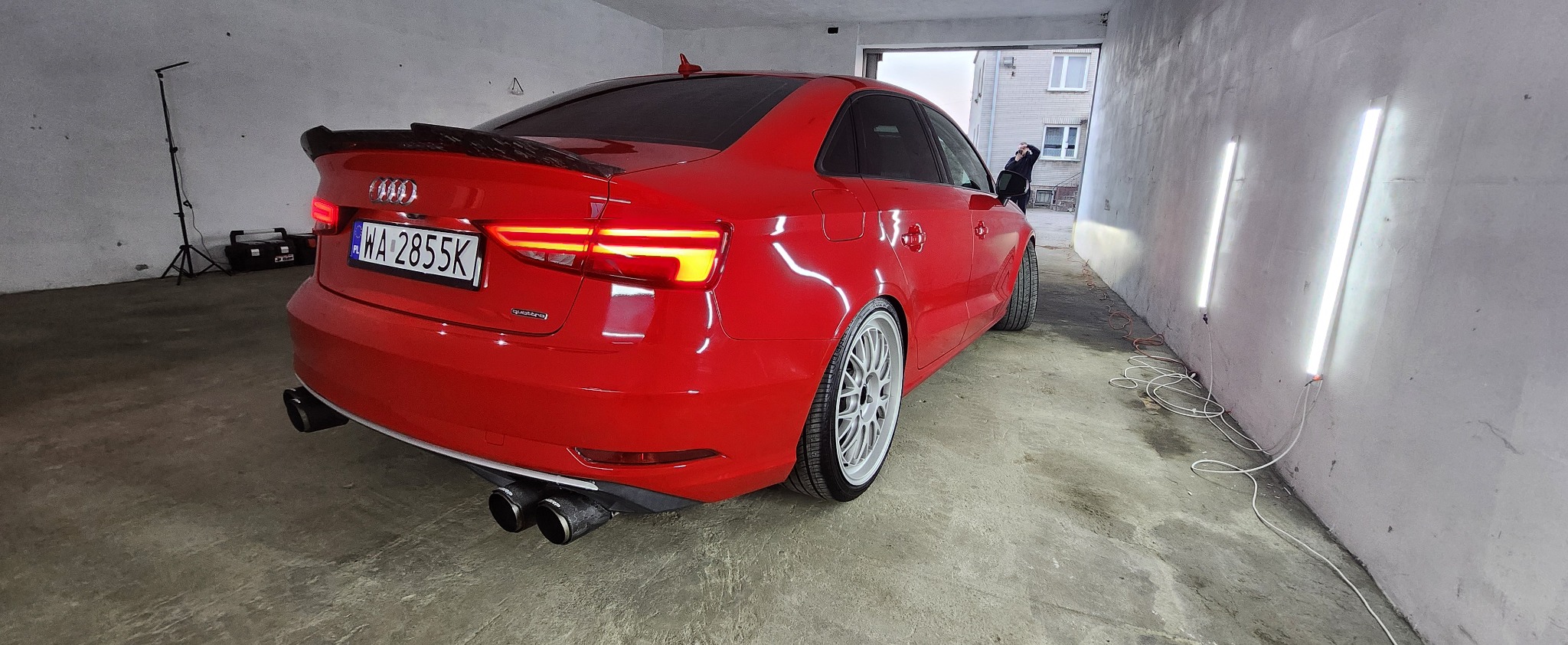 Czerwone Audi A3 po tuningu, z widocznym spojlerem, felgami i podwójnym wydechem, sfotografowane w garażu z oświetleniem LED. Tablica rejestracyjna WA 2855K.