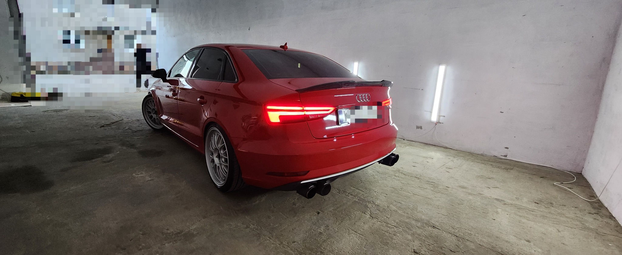 Czerwone Audi A3 z tuningowanym spojlerem i czterema końcówkami wydechu, sfotografowane w warsztacie pod kątem, z włączonymi tylnymi światłami.