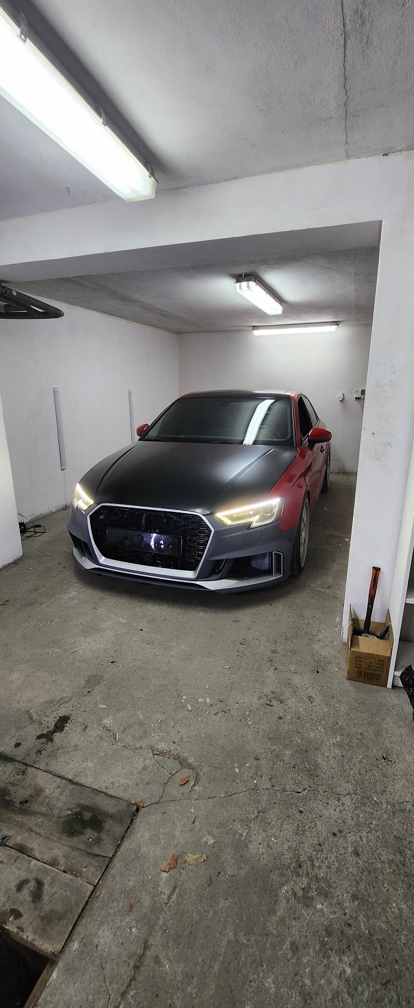 Czerwony Audi A3 z czarną maską w garażu. Widoczne narzędzia i częściowo zdemontowany zderzak. Oświetlenie jarzeniowe. Elementy tuningu samochodowego.