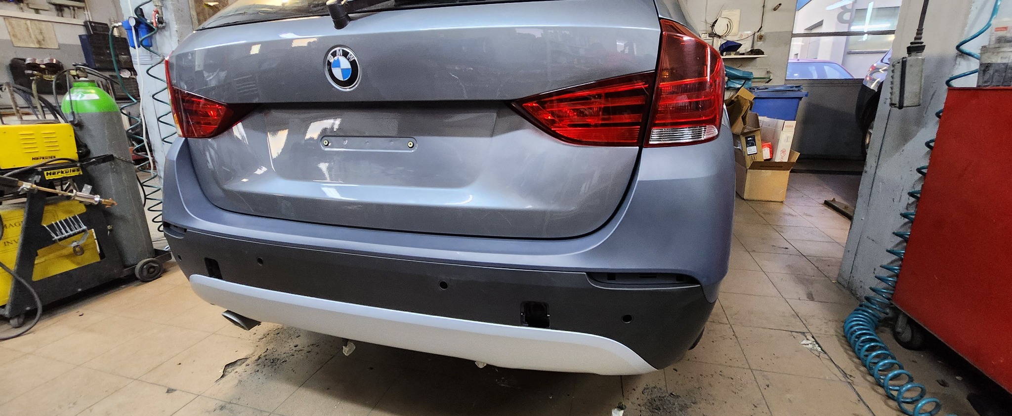 Szary samochód BMW w warsztacie, widok z tyłu, z widocznym spawarką i butlą z gazem w tle. Detale karoserii i wyposażenie warsztatu.