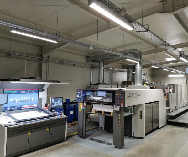 Nowoczesna maszyna offsetowa Lithrone w hali drukarni, z kontrolnym stanowiskiem komputerowym i zapasem papieru. Oświetlenie sufitowe, przemysłowe otoczenie.