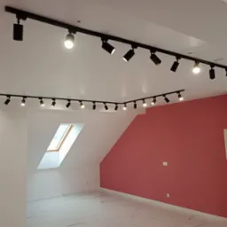 Nowoczesne oświetlenie szynowe z reflektorami w pokoju na poddaszu, z białymi ścianami i jedną ścianą w kolorze brudnego różu. Widoczne okno dachowe.