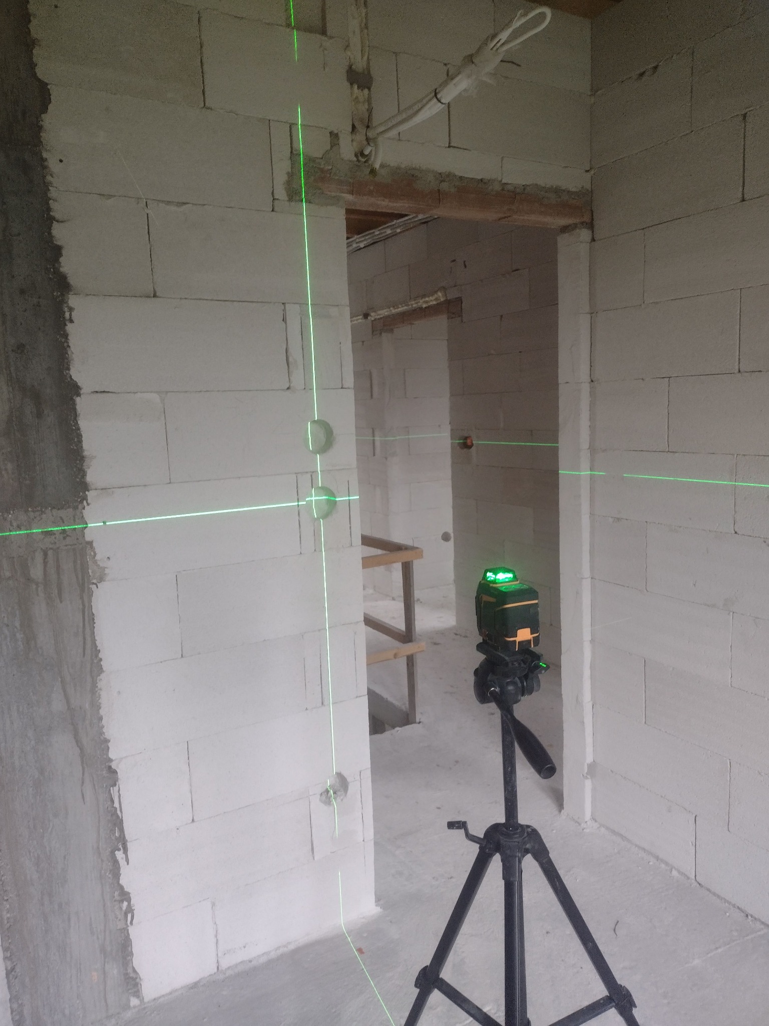 Laser krzyżowy na statywie wyznacza pion i poziom na ścianie z betonu komórkowego z wywierconymi otworami. Widoczne przewody elektryczne.