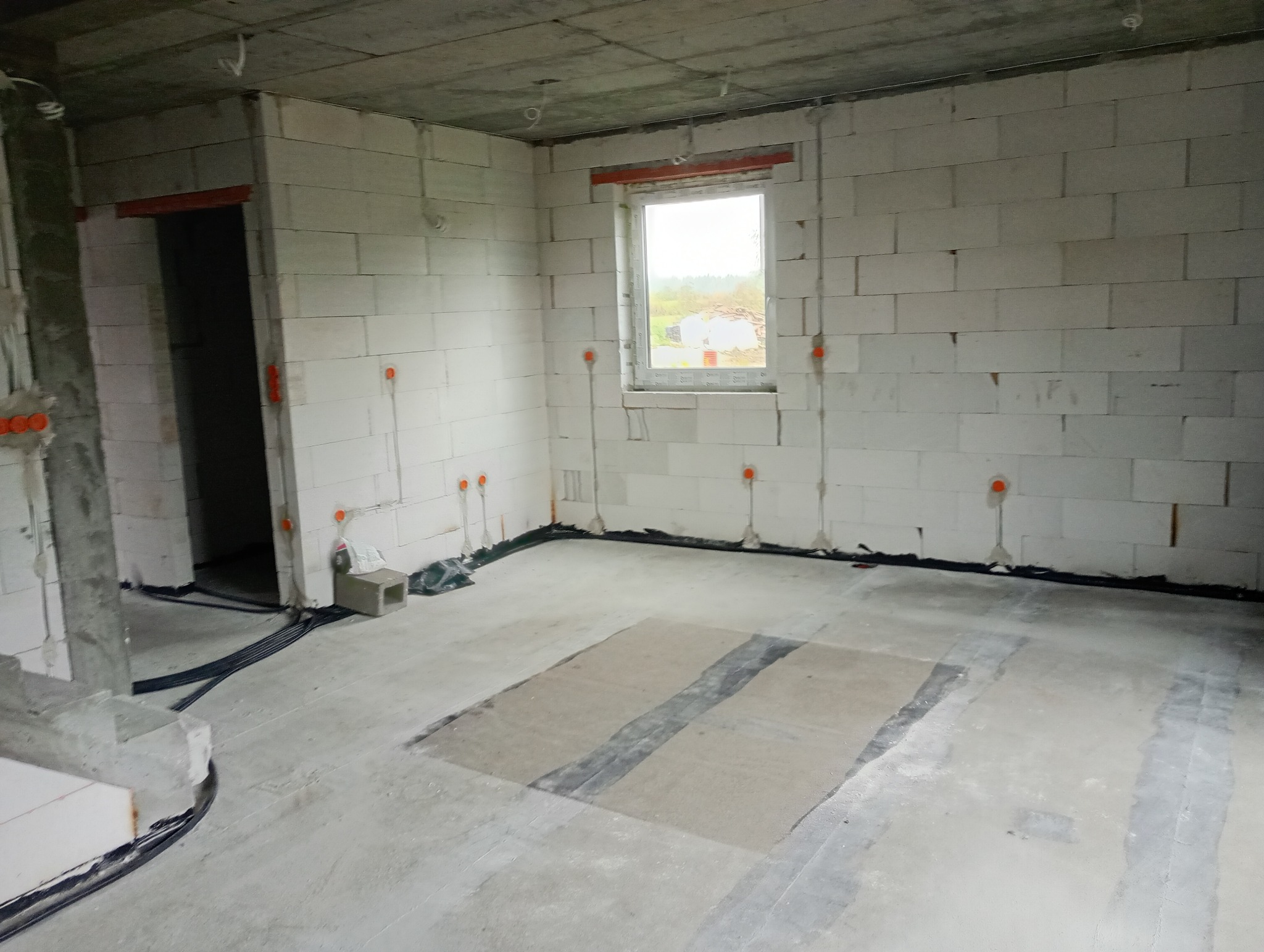 Surowy stan wnętrza z instalacją elektryczną: widoczne kable w peszlach, puszki podtynkowe na ścianach z betonu komórkowego. Okno z widokiem na zewnątrz.