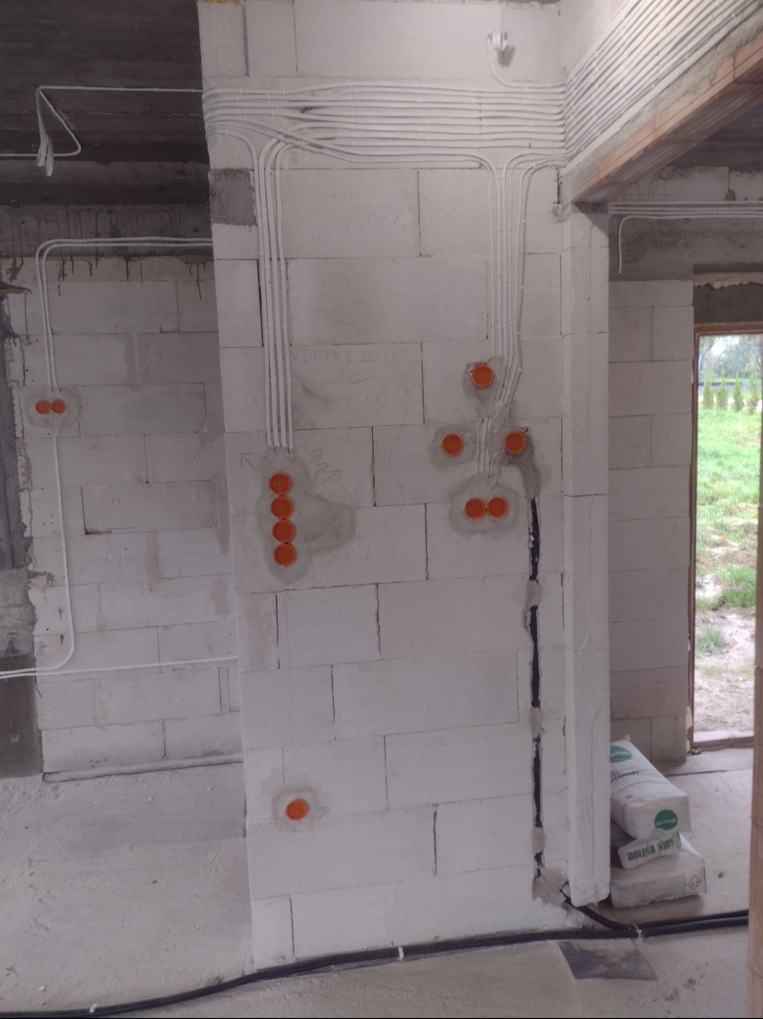 Białe ściany z pustaków z widoczną, częściowo zamontowaną instalacją elektryczną w peszlach i puszkami. Widoczne worki z zaprawą. Surowy stan wykończenia.