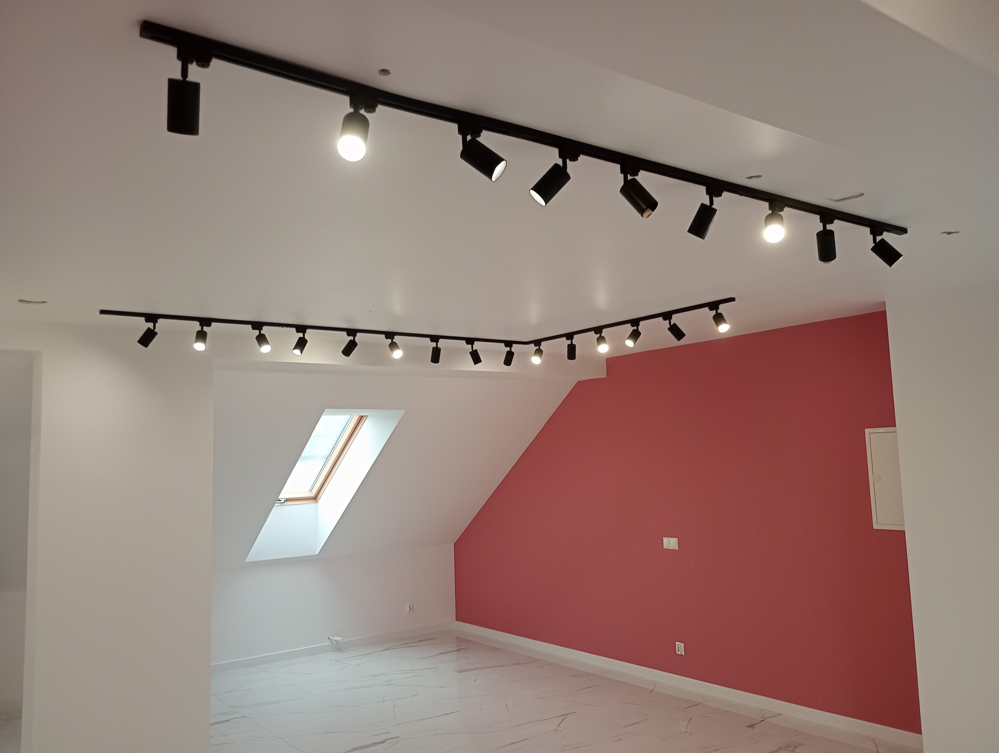 Nowoczesne oświetlenie szynowe z reflektorami w pokoju na poddaszu, z białymi ścianami i jedną ścianą w kolorze brudnego różu. Widoczne okno dachowe.