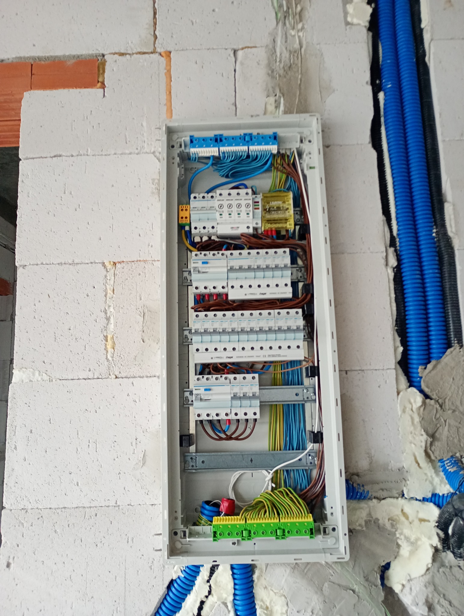 Rozdzielnia elektryczna w trakcie montażu w surowym wnętrzu. Widoczne kable, bezpieczniki i rury instalacyjne. Ściany z białego bloczka.