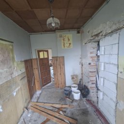 Emil Uzarowski - Wnętrze w trakcie remontu: odsłonięte ściany, drewniane panele, wiadra i narzędzia. Widoczny fragment sufitu z lampą. Prace przygotowawcze do wykończenia.