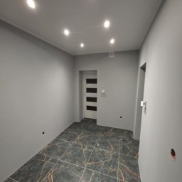 Emil Uzarowski - Wykończone wnętrze w Wschowie: szare ściany, podłoga z płytek imitujących marmur z złotymi żyłami, białe drzwi z czarnymi paskami i oświetlenie punktowe.