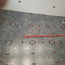 Emil Uzarowski - Układanie płytek imitujących marmur na ścianie, z użyciem poziomicy i klipsów do wyrównywania. Widoczne otwory w suficie i pojemnik z narzędziami.