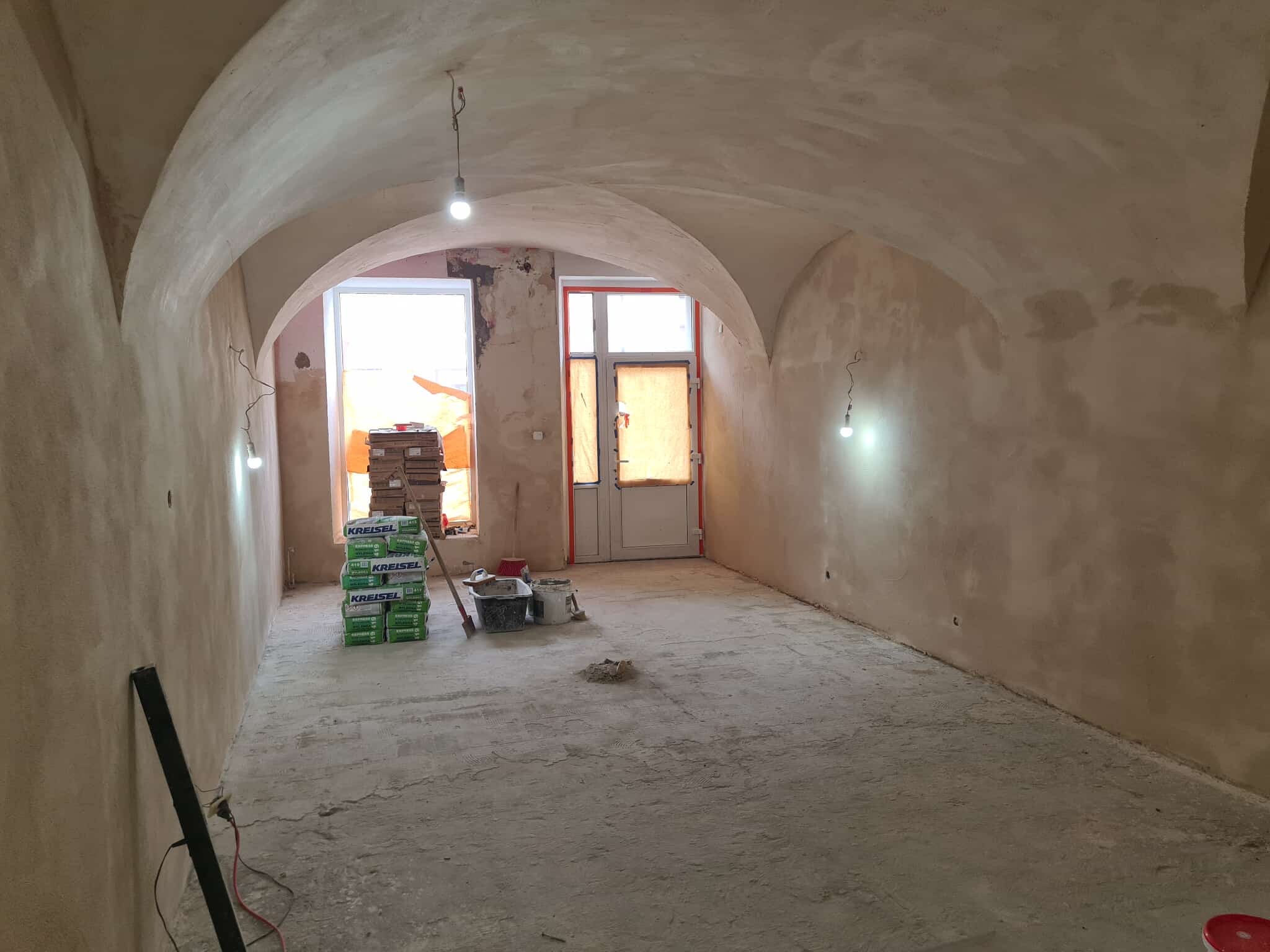 Wnętrze w trakcie prac wykończeniowych: ściany pokryte gładzią, sufit łukowy, worki z zaprawą Kreisel, narzędzia murarskie. Widoczne niedoskonałości i surowy stan.