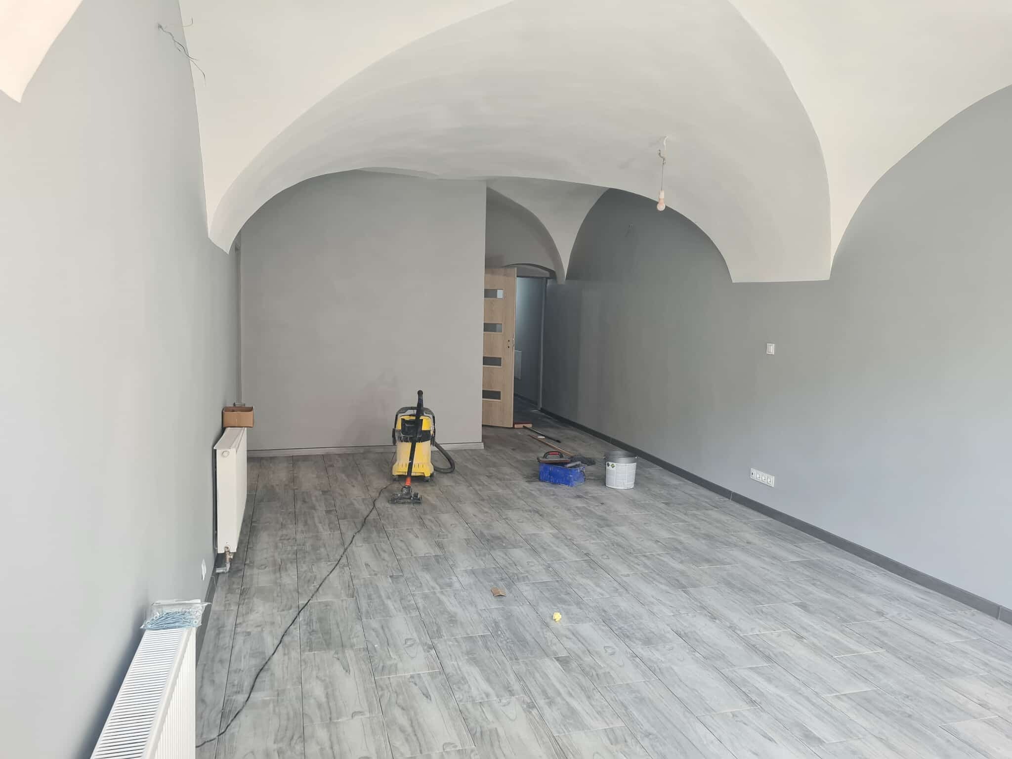 Szare wnętrze po remoncie z łukowym sufitem. Widoczny odkurzacz przemysłowy, grzejniki i podłoga imitująca drewno. Ściany pomalowane na szaro.