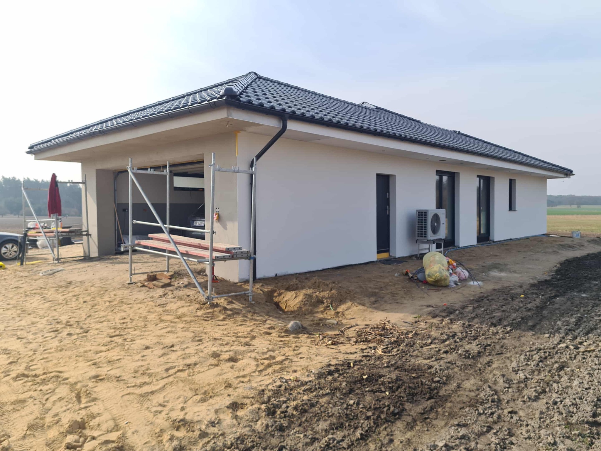 Parterowy dom z szarą dachówką po remoncie elewacji, z widocznym rusztowaniem i elementami wykończeniowymi na zewnątrz. Widoczne okna i drzwi w kolorze czarnym.