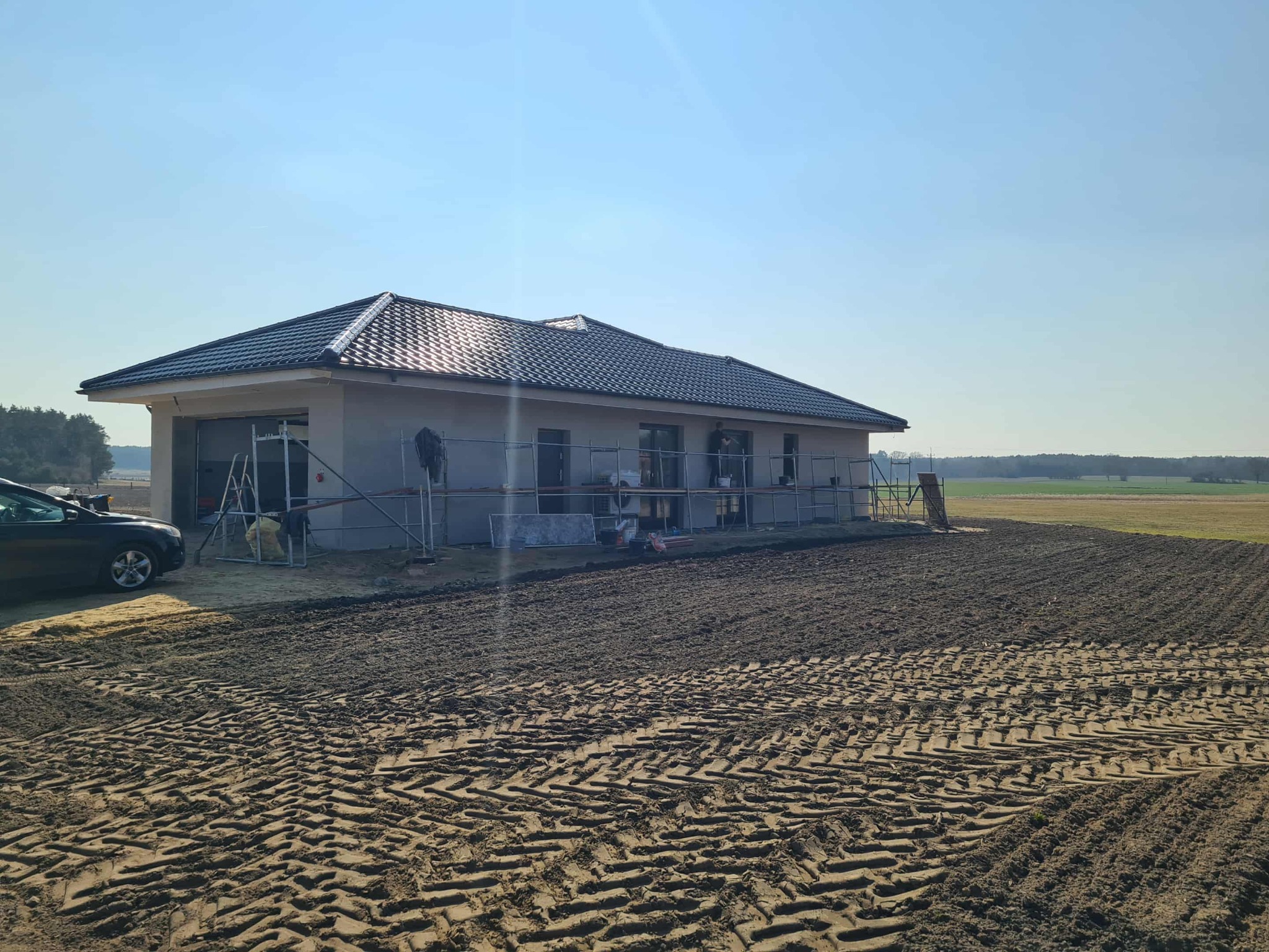 Dom jednorodzinny w trakcie budowy, z widocznymi rusztowaniami i przygotowanym terenem wokół. Szary budynek z ciemnym dachem na tle błękitnego nieba i zieleni.