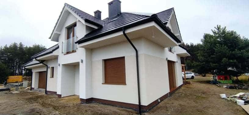 Nowoczesny, dwukondygnacyjny dom z białymi ścianami i ciemną dachówką, brązowe rolety okienne, widok z zewnątrz, teren wokół domu w trakcie zagospodarowywania.