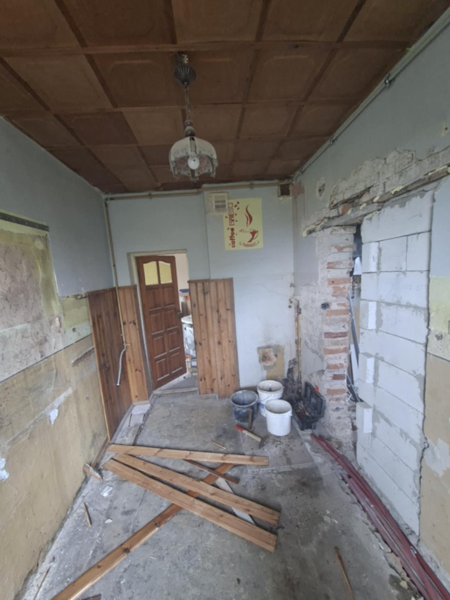 Wnętrze w trakcie remontu: odsłonięte ściany, drewniane panele, wiadra i narzędzia. Widoczny fragment sufitu z lampą. Prace przygotowawcze do wykończenia.