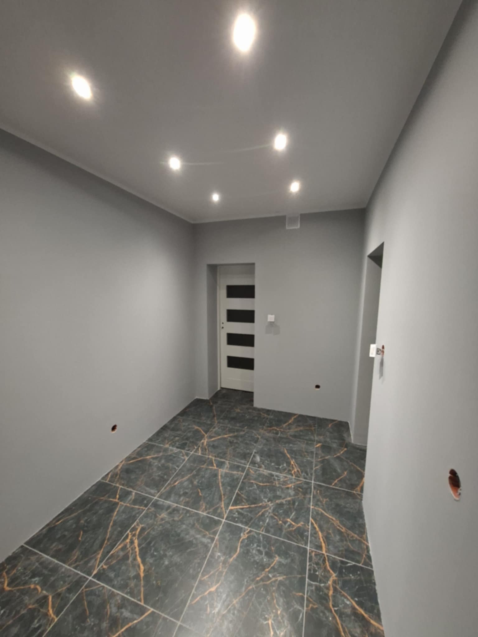 Wykończone wnętrze w Wschowie: szare ściany, podłoga z płytek imitujących marmur z złotymi żyłami, białe drzwi z czarnymi paskami i oświetlenie punktowe.