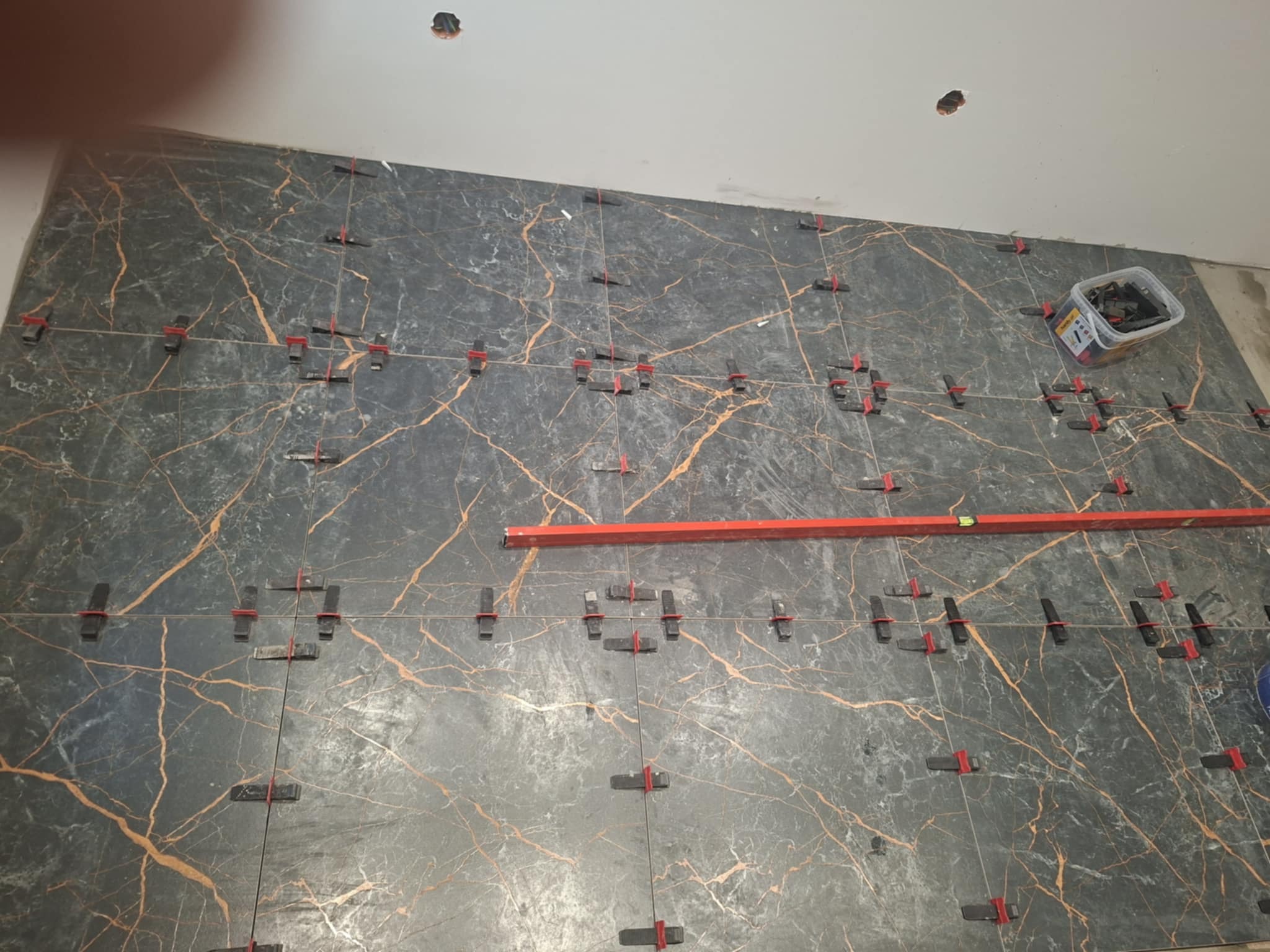 Układanie płytek imitujących marmur na ścianie, z użyciem poziomicy i klipsów do wyrównywania. Widoczne otwory w suficie i pojemnik z narzędziami.